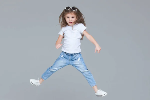 Dívka školačka v modrých džínách, bílé tričko a sluneční brýle je skákání se zvednutýma rukama izolované na šedém pozadí — Stock fotografie