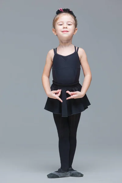 Beautiful little girl gymnast in black sportswear is going to become ballet dancer and shows exercises on grey background — Stock Photo, Image