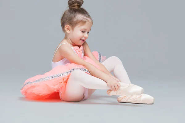 Bonito encantador bailarina menina em tutu rosa está sentado no chão tentando colocar em sapatos de balé — Fotografia de Stock