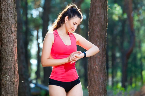 Mulher bonita corredor olha para o seu rastreador de fitness se preparando para uma corrida na floresta de verão — Fotografia de Stock