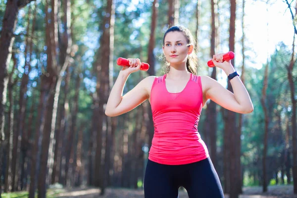 Menina atraente esportiva trabalhando com halteres, treinando na floresta. Fitness, esporte, conceito de estilo de vida — Fotografia de Stock