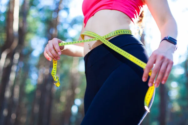 Foto de uma jovem mulher em forma e saudável medindo sua cintura com uma fita métrica controlando sua perda de peso durante o treinamento na floresta — Fotografia de Stock
