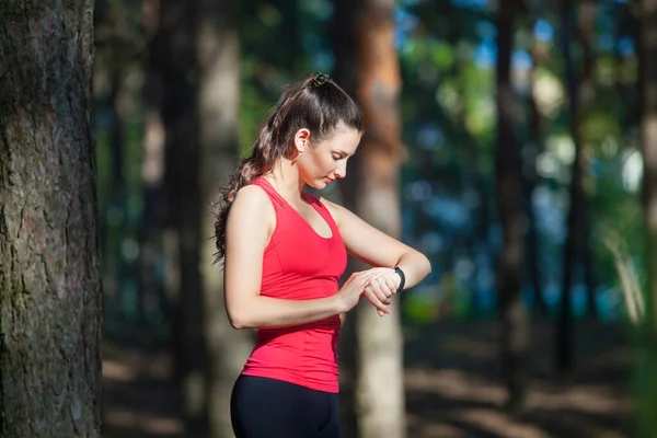 Beautiful woman runner looks at her fitness tracker getting ready for a run in the summer forest — Stock Photo, Image