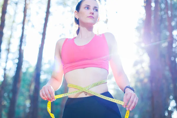 Foto de uma jovem mulher em forma e saudável medindo sua cintura com uma fita métrica controlando sua perda de peso durante o treinamento na floresta — Fotografia de Stock