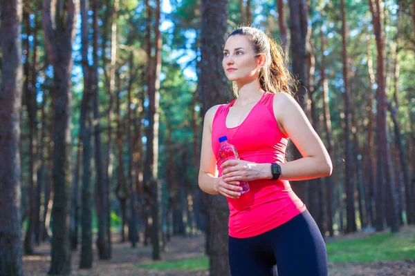 산뜻 하고 날씬 한 젊은 여성, 병에 담긴 생수를 마시며 숲 속에서 달리는 훈련을 멈추는 모습 — 스톡 사진