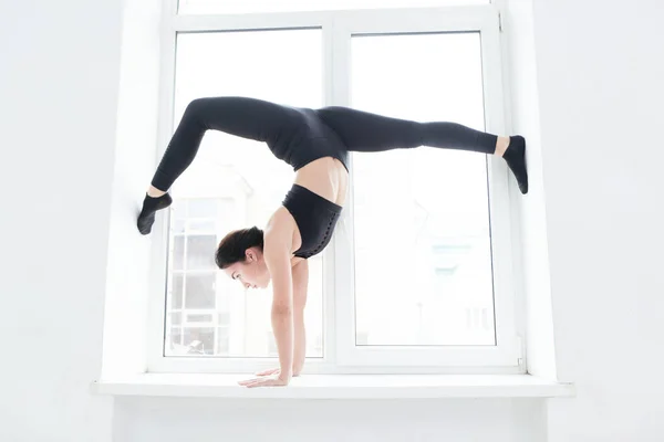 Young woman practicing yoga, standing in handstand exercise on the windowsill, working out wearing black sportswear — Stock Photo, Image