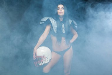 Amerikan futbolu. Siyah arka planda izole edilmiş dumanlı bir kaskla poz veren seksi rugby futbolcusu üniforması giyen genç esmer kız ön planda.