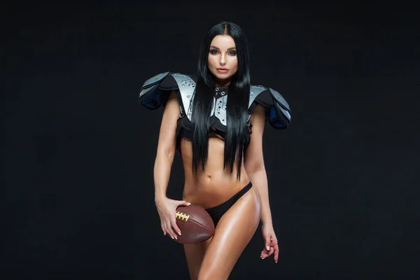 Американський футбол. Молода спортивна брюнетка, одягнена в сексуальну форму регбійного футболіста, позує з м'ячем, ізольованим на чорному тлі. — стокове фото