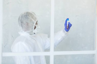Kimyasal koruma giysisi içindeki doktor pencerenin arkasında duruyor ve elinde virüsün salgını durdurmak için COVID, COVID-19 ve koronavirüs üzerinde kan örneği tutuyor.