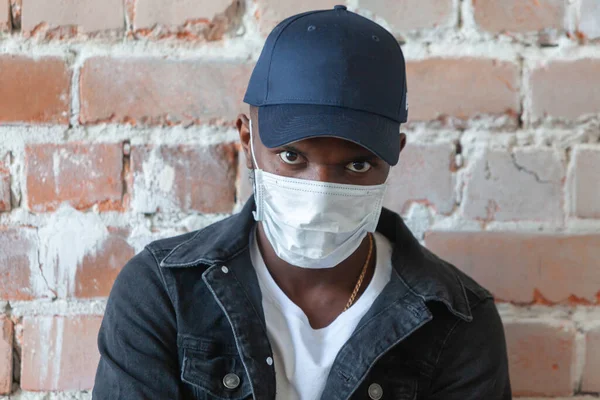 Молодой афроамериканец в медицинской маске стоит в шапке со скрещенными руками в шоке от коронавируса 2019 года на фоне разрушенной стены из красного кирпича — стоковое фото