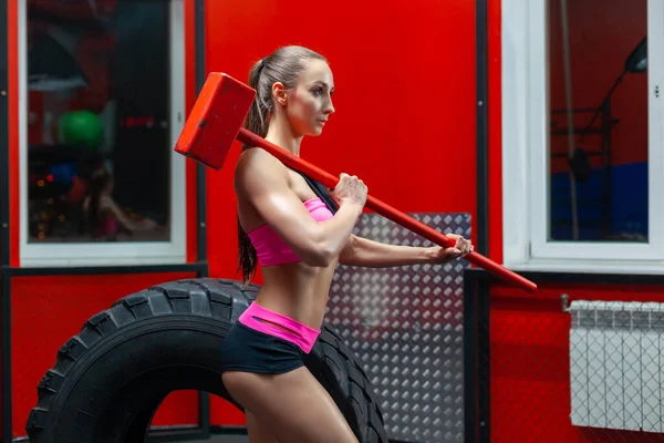 Vista lateral de uma mulher fitness muscular posando perto de pneu com marreta em seu ombro em um ginásio moderno — Fotografia de Stock