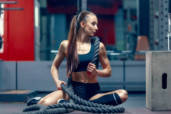 En attraktiv ung muskulös flicka sitter trött på golvet efter slaget rep motion i ett gym — Stockfoto