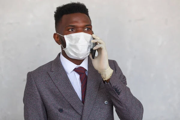 COVID-19. Красивый африканский черный мужчина в костюме использует медицинскую маску и держит свой мобильный телефон рукой в перчатках. Меры по предотвращению коронавирусной инфекции и развитию пандемии — стоковое фото