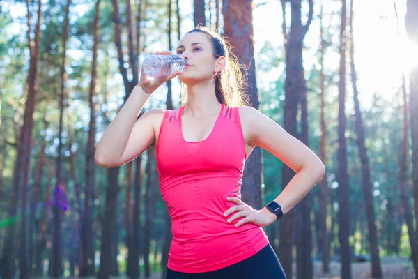 산뜻 하고 날씬 한 젊은 여성, 병에 담긴 생수를 마시며 숲 속에서 달리는 훈련을 멈추는 모습 — 스톡 사진