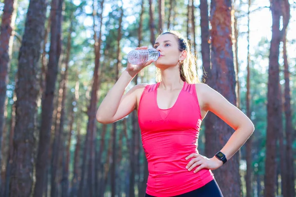 Přizpůsobit štíhlé mladé ženy ve sportovním oblečení pití balené vody, když se zastaví trénink běh v lese — Stock fotografie