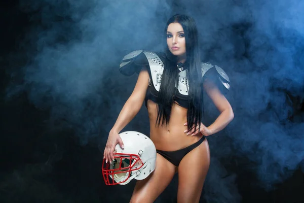 American football. Front view of young sporty brunette wearing sexy uniform of rugby football player posing with helmet in smoke isolated on black background — Stock Photo, Image