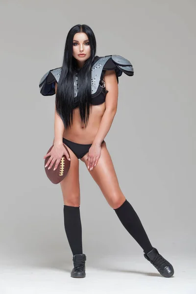 Belle brune sexy joueuse de football américaine en uniforme posant avec une balle isolée sur fond gris — Photo