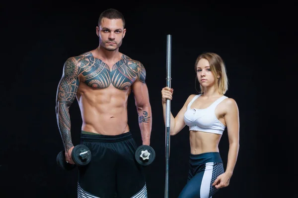 Muscular Fitness hombre con un tatuaje y una mujer deportiva posando sobre un fondo negro con mancuernas y barra —  Fotos de Stock