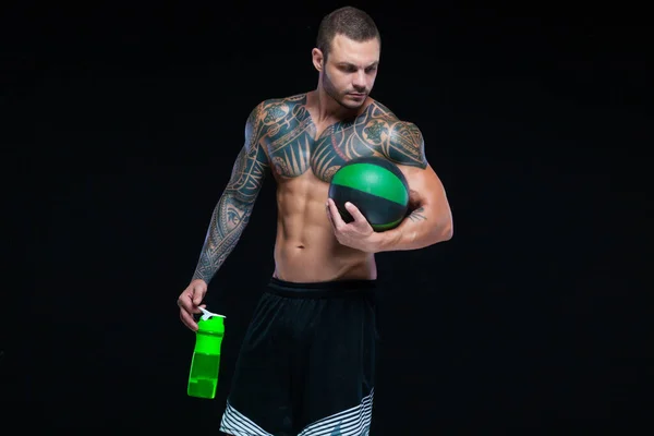 Muscular hombre culturista con tatuajes. Hombre posando sobre un fondo negro con botella de agua y bola — Foto de Stock