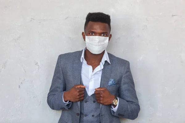 COVID-19. Уверенный молодой африканский мужчина носит защитную медицинскую маску для лица, чтобы предотвратить заражение коронавирусом и пандемию, стоящую в сером костюме у бетонной стены — стоковое фото