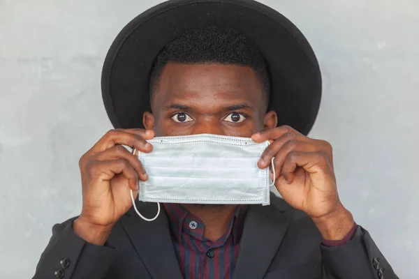 COVID, COVID-19, coronavirus, pandemic. Young African man is going to put on a protective medical face mask to prevent infection with coronavirus and pandemic standing in a suit and black hat against — Stock Photo, Image