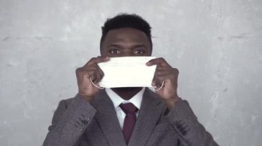 Beton duvara karşı takım elbiseli siyahi bir adamın, tıbbi bir maske takıp yüzüne yerleştirdiği ön görüntüyü kapatın. Koronavirüs, salgın, salgın, COVID-19, grip, soğuk algınlığı.