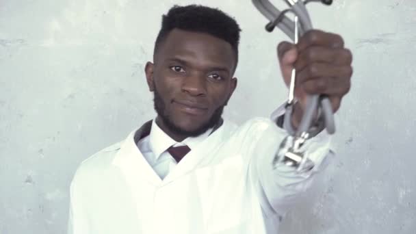 Vista frontal de un médico negro con una bata blanca de laboratorio de pie contra una pared de hormigón, que levanta la mano con un estetoscopio delante de él y luego lo baja. Coronavirus, epidemia, pandemia — Vídeo de stock