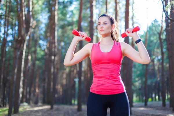 Chica atractiva deportiva haciendo ejercicio con pesas, entrenando en el bosque. Fitness, deporte, concepto de estilo de vida — Foto de Stock