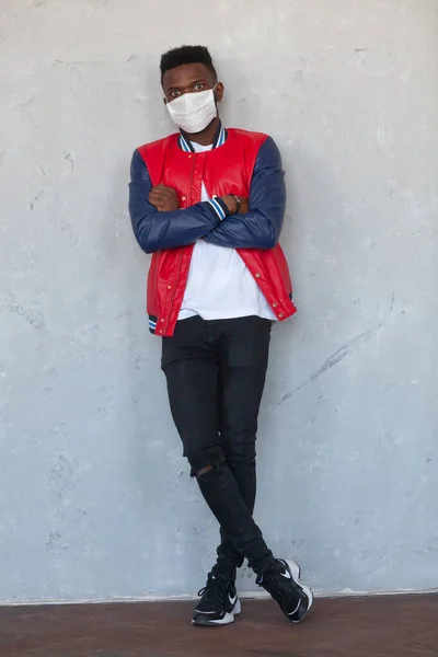 2020 Coronavirus salgınından korunmak için maske takan kolları ve bacakları çaprazlanmış gri beton duvara karşı duran kırmızı ceketli ve kot pantolonlu yakışıklı Afrikalı adamın tam boy portresi. — Stok fotoğraf
