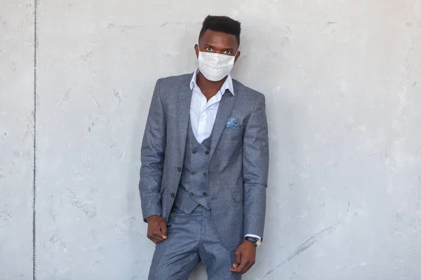 COVID-19. Koronavirüslü enfeksiyonu ve salgını önlemek için koruyucu bir tıbbi maske takan kendinden emin genç Afrikalı bir adam gri bir giysinin içinde beton bir duvara karşı duruyor. — Stok fotoğraf