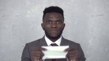 Siyahi bir adamın beton bir duvara karşı takım elbise giymesi, tıbbi bir maskeyi kaldırıp yüzüne dayaması ve sonra da indirmesi. Koronavirüs, salgın, salgın, COVID-19, grip, soğuk algınlığı.