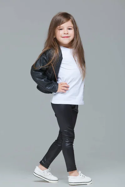 Niña con el pelo largo en chaqueta de cuero negro y leggings está de pie en el estudio sobre fondo gris — Foto de Stock