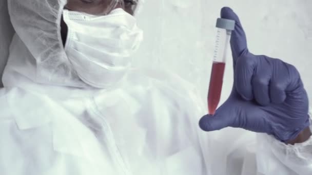 Close-up van het bloedmonster tegen het gezicht gehouden door een mannelijke laboratoriummedewerker in een chemisch beschermingspak, die het onderzoekt en goedkeurend knikt achter het glas in het laboratorium. Coronavirus, epidemie — Stockvideo