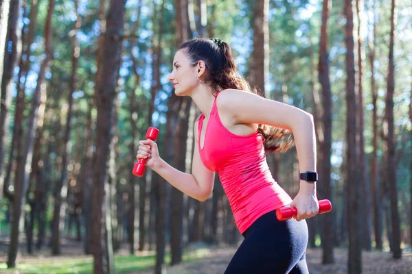 Vue latérale d'une jolie fille sportive s'entraînant avec des haltères, s'entraînant dans la forêt. Fitness, sport, concept lifestyle — Photo