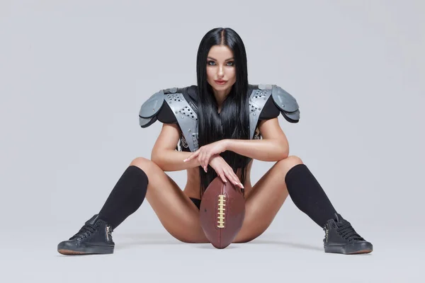 Beautiful sporty brunette dressed in bikini and American football uniforms posing sitting on the floor with a ball between her legs isolated on grey background — Stock Photo, Image