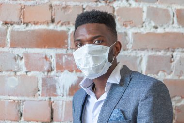 Kırmızı tuğlalı duvara karşı takım elbiseli Afrikalı genç bir siyahi adam Coronavirus salgınından korkuyor ve COVID-19 enfeksiyonuna yakalanmamak için tıbbi maske takıyor.