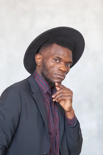 Siyah takım elbiseli, şapkalı, gri beton duvara bakan Afrikalı genç bir adam. — Stok fotoğraf