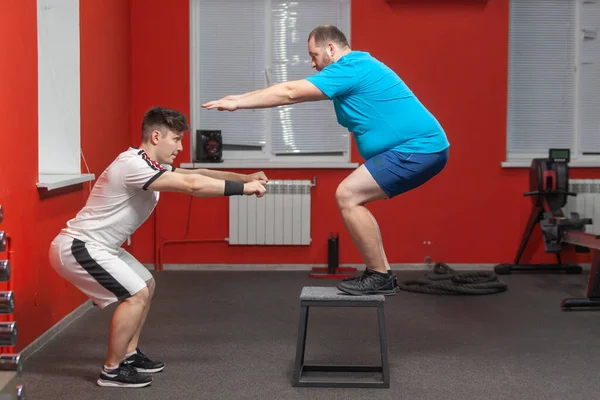 Ansioso homem com excesso de peso está fazendo saltar no ginásio sob a orientação de um personal trainer — Fotografia de Stock