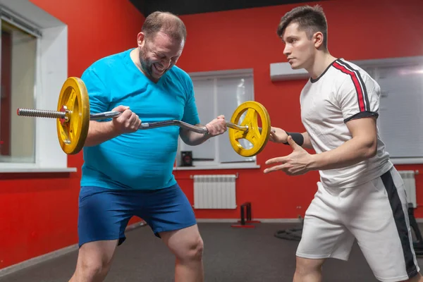 健身房里的一个胖子在私人教练的指导下，在他的能力范围内进行杠铃举重练习。体重超标 — 图库照片