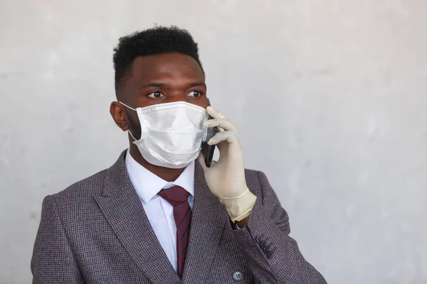 COVID-19. Красивый африканский черный мужчина в костюме использует медицинскую маску и держит свой мобильный телефон рукой в перчатках . — стоковое фото