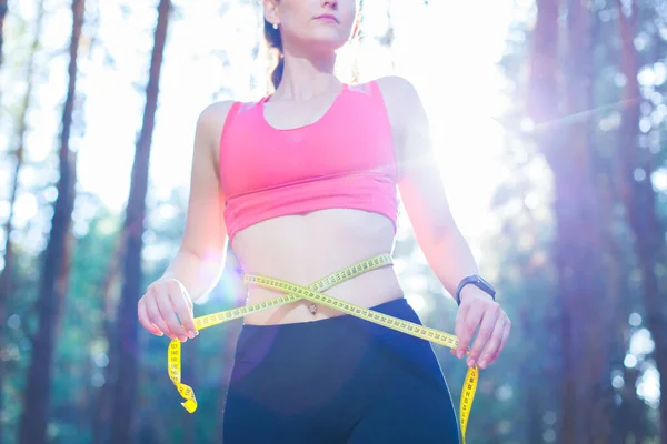 Foto de uma jovem mulher em forma e saudável medindo sua cintura com uma fita métrica controlando sua perda de peso durante o treinamento na floresta — Fotografia de Stock