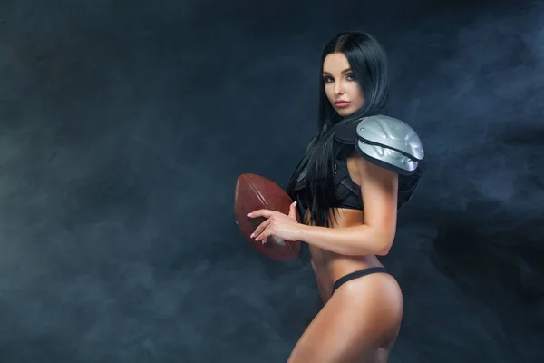 American football. Young sporty brunette wearing sexy uniform of rugby football player posing with ball in smoke isolated on black background — Stock Photo, Image