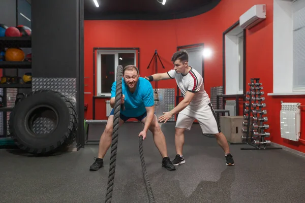 El joven gordo está realizando ejercicio de cuerda de batalla en el gimnasio de fitness con la última de sus fuerzas bajo el control de su entrenador personal. Peso excesivo —  Fotos de Stock