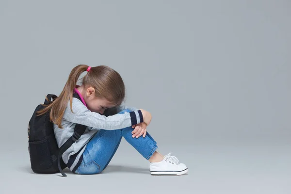 Gadis sekolah dasar dalam jins dan jaket seragam memakai ransel duduk lelah setelah kelas. Terisolasi di latar belakang abu-abu — Stok Foto