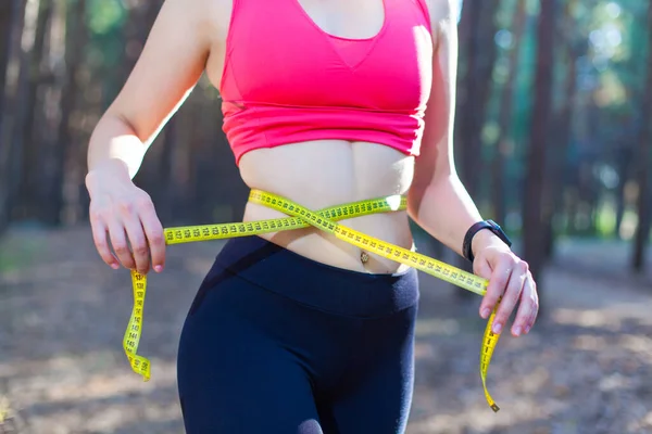 Foto de uma jovem mulher em forma e saudável medindo sua cintura com uma fita métrica controlando sua perda de peso durante o treinamento na floresta — Fotografia de Stock