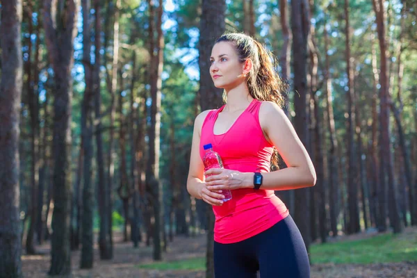 S'adapter jeune femme mince en vêtements de sport boire de l'eau embouteillée comme elle arrête une course d'entraînement dans la forêt — Photo