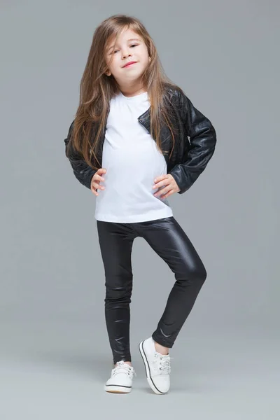 Niña con el pelo largo en chaqueta de cuero negro y leggings está de pie en el estudio sobre fondo gris — Foto de Stock