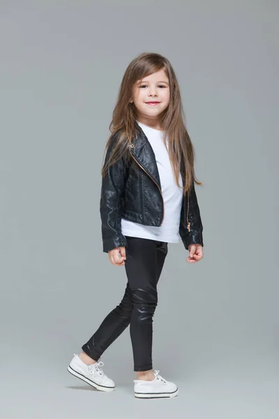 Niña con el pelo largo en chaqueta de cuero negro y leggings está de pie en el estudio sobre fondo gris — Foto de Stock