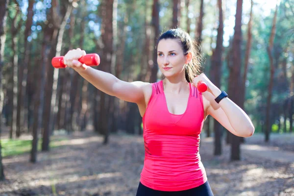 Menina atraente esportiva trabalhando com halteres, treinando na floresta. Fitness, esporte, conceito de estilo de vida — Fotografia de Stock