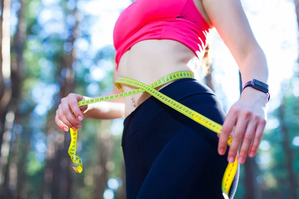 Photo d'une jeune femme en forme et en bonne santé mesurant sa taille avec un ruban à mesurer contrôlant sa perte de poids tout en s'entraînant dans la forêt — Photo